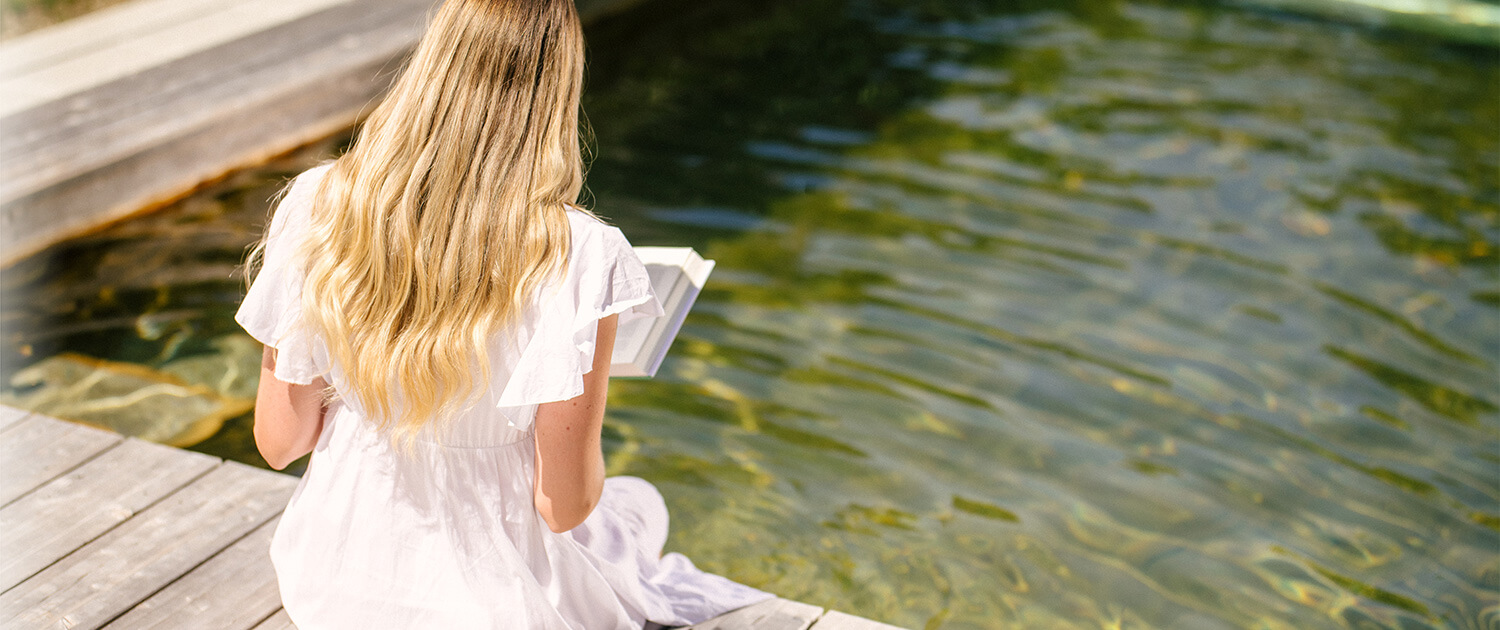 Dame liest ein Buch am Holc Naturpool und hält die Beine ins Wasser.